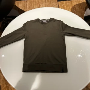 Mörkgrön sweatshirt från Calvin Klein - Snygg mörkgrön sweatshirt från Calvin Klein med diskret logotyp broderad framtill. Tröjan har rund hals, långa ärmar och ribbade muddar vid ärmslut och nederkant. Perfekt för dig som gillar stilrena och enkla plagg med en touch av designer.