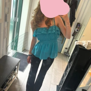 Turkos volangblus med smock - Säljer en turkos blus med volanger upptill och smock i midjan. Blusen är ärmlös med breda axelband och har en luftig, flowy passform. Perfekt för dig som gillar färg och romantiska detaljer.