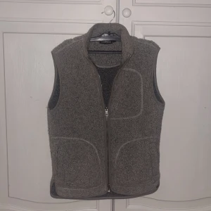 Grå fleeceväst från J.Lindeberg - Säljer en grå fleeceväst från J.Lindeberg med hög krage och hel dragkedja framtill. Västen har ett mysigt teddy-material och praktiska fickor på sidorna. Perfekt att slänga på över en t-shirt för extra värme och stil. Storlek S 