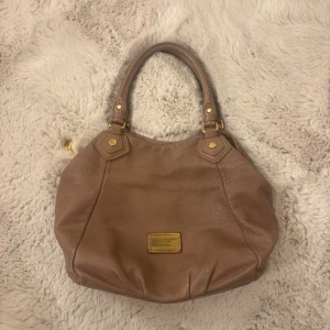Brun handväska från Marc Jacobs - Snygg handväska från Marc Jacobs i en mjuk brun nyans med guldfärgade metalldetaljer och logoplatta framtill. Väskan är tillverkad i äkta skinn och har dubbla handtag med dekorativa nitar och spännen. Perfekt storlek för att få plats med allt du behöver, t.ex. skolan. Vintage och modellen säljs ej längre. 