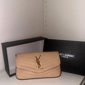 Beige quiltad YSL handväska i skinn - Supersnygg oanvänd beige handväska från Saint Laurent med quiltad design och guldigt YSL-emblem framtill. Väskan är i skinn och har en avtagbar guldkedja samt flera fack för kort och småsaker. Perfekt för dig som vill ha en lyxig och stilren accessoar.