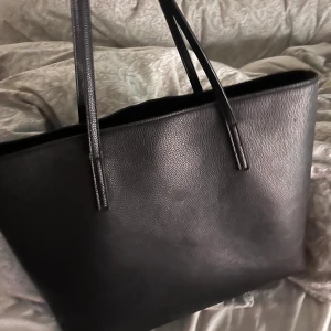 Svart shoppingväska i skinn - Stilren svart shoppingväska med två långa handtag, perfekt för att bära allt du behöver. Väskan är tillverkad i slätt skinn och har en rymlig insida utan extra fack. Enkel och klassisk design som funkar till många olika outfits.