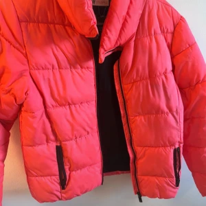 Rosa pufferjacka med dragkedja - Säljer en riktigt snygg rosa pufferjacka med hög krage och svarta dragkedjor på fickorna. Jackan har quiltad design och är perfekt för kalla dagar.🏂🎿 Den har en loose passform och är tillverkad i syntetmaterial som håller värmen bra. Snabb leverans 📦 