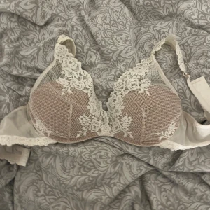 Beige spetsblus med broderad spets - Säljer en beige blus med vacker vit spets och broderade blommor. Toppen har en feminin design med spetsdetaljer längs kuporna och axelbanden. Perfekt för dig som gillar romantisk och elegant stil.