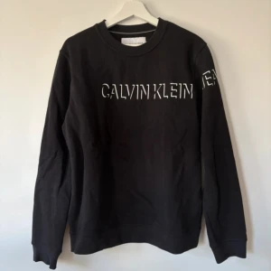 Svart sweatshirt från Calvin Klein Jeans - Svart sweatshirt från Calvin Klein Jeans med vit logotyp framtill och på ärmen. Tröjan har rund hals, långa ärmar och är tillverkad i mjuk bomull med inslag av polyester. Enkel och stilren design som passar perfekt till jeans eller joggers.