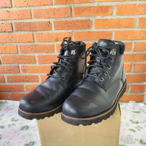 Svarta UGG boots i skinn - Säljer ett par svarta boots från UGG med robust snörning och grov platt sula. Skorna är tillverkade i skinn med detaljer i mocka vid skaftet och har metallöglor för snörningen. UGG-logga bak på hälen. Perfekt för dig som gillar stilrena och tåliga boots.