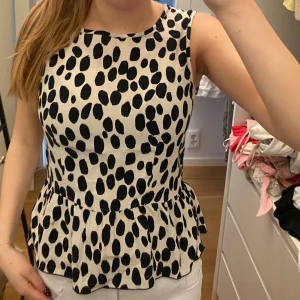 Svartvit prickig peplum blus - Säljer en svartvit blus med peplumvolang och stora svarta prickar på vit botten. Toppen är ärmlös, har rund halsringning och dragkedja i ryggen. Perfekt för dig som gillar lekfulla mönster och en feminin siluett. Den har en fläck men den syns inte när tröjan sitter på