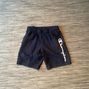 Mörkblå Champion shorts i bomull - Snygga mörkblå shorts från Champion med vit logga längs benet. Modellen har elastisk midja och är tillverkad i mjuk bomull och polyester, vilket gör dem sköna att ha på sig. Perfekta för chill eller träning, klassisk stil med normal passform.