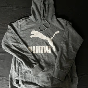 Grå hoodie från Puma med logga - Säljer en grå hoodie från Puma i storlek M. Tröjan har stor vit Puma-logga framtill, huva med snörning och ribbade muddar vid ärmslut och nederkant. Perfekt för chill dagar eller när du vill ha en sportig look. Notera att de ena snöret saknar plasten och att det har gått upp lite vid snöret 👕