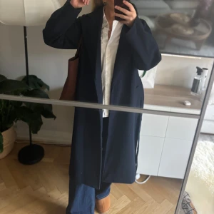 Mörkblå kappa med bälte - Säljer en stilren mörkblå kappa med bälte i midjan och klassisk krage. Modellen är lång och har en loose passform med långa ärmar. Perfekt för dig som gillar minimalistisk och clean look. Materialet känns mjukt och har en ullig känsla.