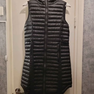 Svart lång dunväst från Everest - Säljer en svart, lång dunväst från Everest med huva och horisontella quiltade sömmar. Västen har dragkedja framtill och två fickor på sidorna. Perfekt för lager-på-lager och extra värme under kyliga dagar.