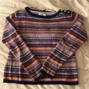 Mönstrad stickad tröja med guldknappar - Färgglad stickad tröja från hampton republic med snyggt randigt mönster i lila, marinblått, gult, rosa och vitt. Rund hals med tre guldiga knappar på axeln som detalj. Långärmad och mjuk, perfekt för dig som gillar retrovibbar och coola färgkombinationer.