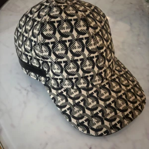 Svartvit keps från Salvatore Ferragamo - Snygg keps från Salvatore Ferragamo med svartvitt grafiskt mönster och diskret läderdetalj baktill. Kepsen har böjd skärm och justerbar rem. Tillverkad i bomull med exklusiv känsla, perfekt för dig som gillar designeraccessoarer.