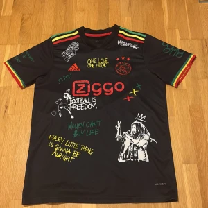 Ajax x Bob Marley svart fotbollströja - Svart Ajax fotbollströja från Adidas med Bob Marley-tema, färgglada ränder i rött, gult och grönt på axlar och ärmslut. Tryck med Ziggo-logga, Ajax-märke, Bob Marley-porträtt och inspirerande texter. Materialet är lätt och andas, perfekt för fotboll eller streetwear. 
