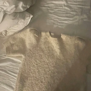 Beige stickad tröja från Indiska - Mysig beige stickad tröja från Indiska med rund hals och korta ärmar. Tröjan har en fluffig och mjuk känsla, perfekt för att hålla dig varm under kyliga dagar. Ribbstickade detaljer vid hals och ärmslut ger en snygg finish.