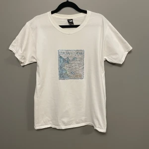 Vit t-shirt med tryck från Bvaldi - Snygg vit t-shirt från Bvaldi i storlek S. Framsidan har ett tryck som liknar ett frimärke med motiv från Italien i blå toner. Klassisk rund hals och korta ärmar. Perfekt för dig som gillar grafiska detaljer och enkel stil.