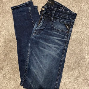 Replay Anbass mörkblå jeans herr - Säljer ett par Replay Anbass jeans i mörkblå tvätt med snygga slitningar och klassiska detaljer. Jeansen har fem fickor, dragkedja och smal passform som sitter riktigt schysst. Perfekta för dig som gillar stilrena och moderna jeans.