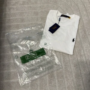 Vit t-shirt från Polo Ralph Lauren - Vit t-shirt från Polo Ralph Lauren med klassisk rund hals och korta ärmar. T-shirten har den ikoniska svarta Polo-loggan broderad på bröstet och är gjord i mjuk bomull. Perfekt basic med stilren design och snygg passform.