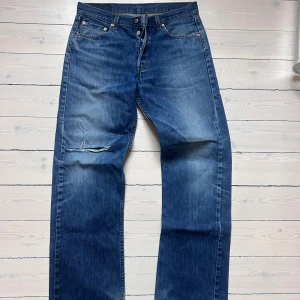 Levi's 501 blå jeans med hål - Klassiska Levi's 501 jeans i blå denim med rak passform och snyggt sliten look. Byxorna har ett stort hål på vänster knä, fem fickor och knappgylf. Perfekta för dig som gillar en avslappnad och vintage-inspirerad stil. W31 L34 modell 187cm 74kg Mad vintage 