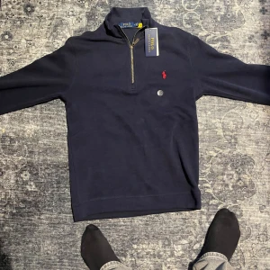 Marinblå half zip tröja Polo Ralph Lauren - Snygg marinblå långärmad tröja från Polo Ralph Lauren med half zip och klassisk röd broderad logga på bröstet. Tröjan har hög krage och är gjord i mjuk bomull, perfekt för lager-på-lager. Passar dig som gillar stilren och tidlös design.