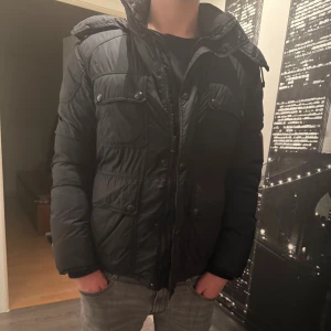 Svart pufferjacka från Belstaff - Svart winter från Belstaff med huva, fyra framfickor med tryckknappar och dragkedja. Materialet är syntet och jackan har en normal passform med långärmade ärmar. Den har en innerficka och varm. Haft den förra vintern men allt är bra. 