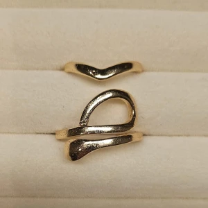 Två guldiga ringar i modern design - Säljer två stilrena ringar i guldton med unik, modern form. Den ena ringen har en mjuk V-form och den andra har en öppen, abstrakt design med svepande linjer. Perfekta för att mixa och matcha för en trendig look.
