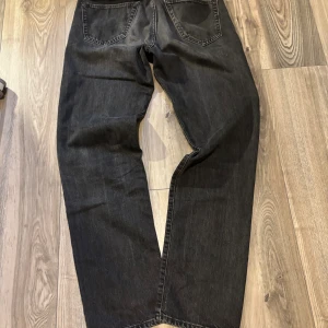 Jack N Jones svarta jeans, storlek 30/34 - Svarta Jack N Jones jeans med klassisk rak passform och fem fickor. Jeansen har en mörk tvätt och tydlig lapp baktill i brunt. Materialet är robust denim och modellen har normal midja. Perfekt för dig som gillar tidlös stil och vill ha ett par jeans som funkar till allt.