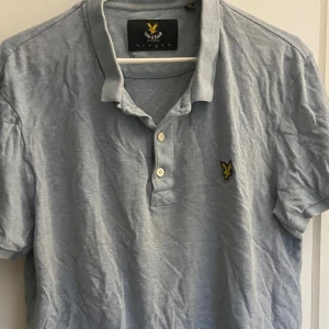 Ljusblå pikétröja från Lyle & Scott - Säljer en ljusblå pikétröja från Lyle & Scott med klassisk krage och två knappar framtill. Tröjan har det ikoniska gula örnloggan broderad på bröstet och är tillverkad i mjuk bomull. Perfekt för en clean och avslappnad stil. L men funkar som M också