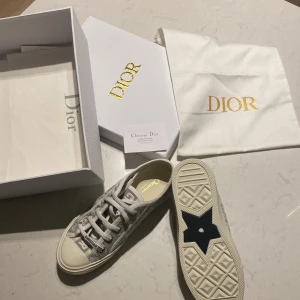 Christian Dior grå sneakers med monogram - Säljer ett par lyxiga Christian Dior sneakers i grått och vitt med tydligt Dior-monogram över hela ovandelen. Skorna har snörning, platt sula med svart stjärndetalj undertill och broderad logga på sidan. Materialet är bomullstyg och gummisula. Perfekt för dig som gillar exklusiv streetstyle.