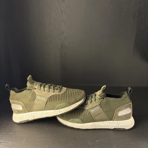 Olivgröna sneakers från BOSS - Snygga olivgröna sneakers från BOSS med stickad ovandel och vita detaljer på sulan. Skorna har snörning, rund tå och en platt sula med mönstrad undersida. Perfekt för dig som gillar stilrena och sportiga skor med modern design.