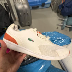 Vita sneakers från Lacoste Sport - Snygga vita sneakers från Lacoste Sport med orange detaljer på häl och plös samt orange sula. Klassisk Lacoste-logga i grönt på sidan. Skorna har rund tå, snörning och är tillverkade i syntet och textil med meshpartier för extra ventilation.