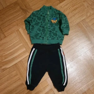 Grön Bamse-kofta och svarta byxor från Lindex - Säljer en grön kofta från Lindex med Bamse-tryck och mönster över hela tröjan. Koftan har dragkedja framtill, ribbade muddar och en söt Bamse-detalj på bröstet. Svarta tillhörande byxor.