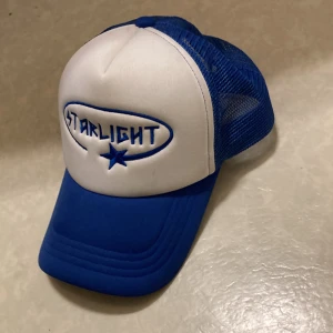 Blå och vit Starlight keps - Snygg truckerkeps från Starlight med vit front och blått broderat Starlight-tryck samt stjärna. Skärmen och mesh-panelerna är blå, vilket ger en fräsch kontrast. Kepsen har klassisk form och justerbar baksida. Perfekt för dig som gillar streetstyle.