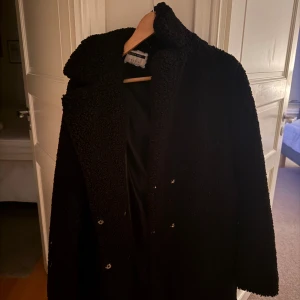 Svart teddyjacka från River Island - Säljer en svart teddyjacka från River Island i storlek 10. Jackan har en fluffig och mjuk yta, bred krage och knäppning framtill. Perfekt för dig som gillar en oversized look och vill ha något som är både stilrent och comfy.