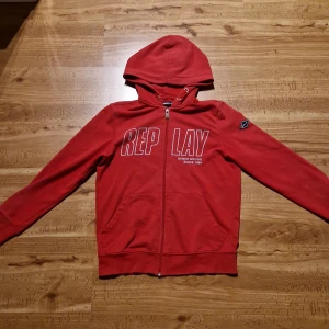 Replay zip hoodie - Röd hoodie från Replay med vit logga på bröstet och patch på ärmen. Tröjan har huva, hel dragkedja och två fickor framtill. Tillverkad i mjuk bomull, perfekt för chill dagar eller när du vill ha en sportig look.