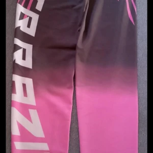 Rosa och svart leggings med tryck - Snygga träningsbyxor i rosa och svart med coolt vitt tryck längs benet. Byxorna har en ombre-effekt från svart till rosa och är tillverkade i ett stretchigt syntetmaterial som passar perfekt för träning eller chill.