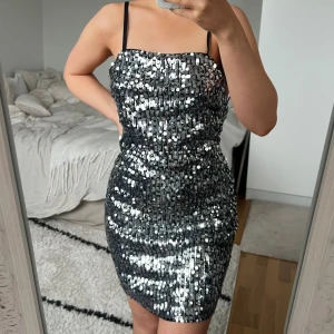 Glittrig silver paljettklänning 🩶 - Säljer en kort, glittrig klänning med silverfärgade paljetter och smala svarta axelband. Klänningen har en tight passform och snygg snörning i ryggen som ger en cool detalj. Perfekt för dig som vill sticka ut med en riktigt festlig look. Storlek S från H&M. Fint skick 🩶🎉