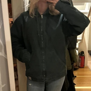 Svart vindjacka från BDG Urban Outfitters - Säljer en mörkgrön jacka från BDG Urban Outfitters i storlek XS. Jackan har huva med snörning, dragkedja framtill och två stora fickor. Materialet är slitstark bomull och insidan är quiltad för extra värme. Perfekt för dig som gillar loose fit och streetstyle.