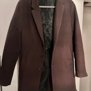 Brun klassisk kappa med knappar - Snygg brun kappa med rak passform och klassisk krage. Kappan har tre svarta knappar framtill och två diskreta fickor på sidorna. Fodrad insida i svart och långärmad design. Perfekt för dig som gillar stilrena och tidlösa plagg.