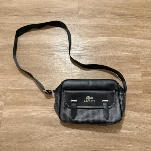 Svart axelväska från Lacoste - Snygg svart axelväska från Lacoste med geometriskt mönster och justerbar rem med logotyp. Väskan har ett lock med två dekorativa remmar och silverfärgade detaljer. Insidan är ljus och har en diskret Lacoste-logga. Perfekt för att bära dina essentials.