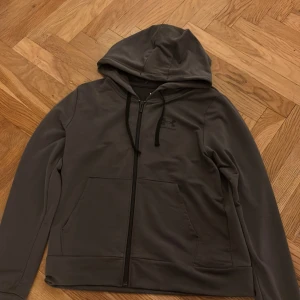 Grå hoodie från Under Armour XS - Snygg grå hoodie från Under Armour i storlek XS. Tröjan har huva med svarta snören, dragkedja framtill och två stora fickor. Loggan är tryckt på bröstet. Passformen är loose och materialet känns mjukt och sportigt, perfekt för chill eller träning.