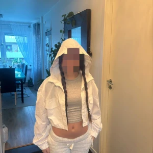 Vit croppad vindjacka med huva - Säljer en fräsch vit croppad vindjacka med huva och snörning i midjan. Jackan har två stora fickor framtill och elastiska muddar vid ärmslut. Perfekt för dig som gillar streetstyle och vill ha en lätt jacka till vår eller sommarkvällar.