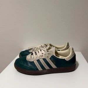 Adidas Samba sneakers mörkgrön - Adidas Samba sneakers i mörkgrönt skinn och mocka med klassiska vita ränder och snörning. Yttersulan är brun och insidan är ljusbeige. Ikonisk retrostil med rund tå och platt sula, perfekt för dig som gillar streetwear och sportig vibe.