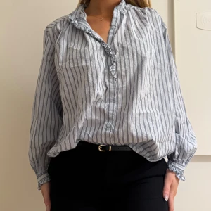 Randig blus  - Luftig och snygg blus i vitt med tunna blå ränder. Skjortan har volangdetalj längs knäppningen, långa ärmar med resår vid ärmslut och en relaxed passform. Perfekt för en chill och stilren look.