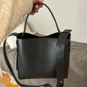 Svart axelväska  - Stilren svart axelväska. Väskan har både handtag och justerbar axelrem, samt ett rymligt huvudfack med innerficka och magnetlås. Perfekt för dig som gillar minimalistisk och tidlös design.