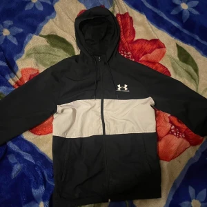 Svart och vit vindjacka Under Armour - Under Armour vindjacka i svart och vitt med huva och dragkedja framtill. Jackan har en clean design med logga på bröstet och praktiska fickor. Perfekt för dig som gillar sportig stil och vill ha något lätt och snyggt att slänga på dig.