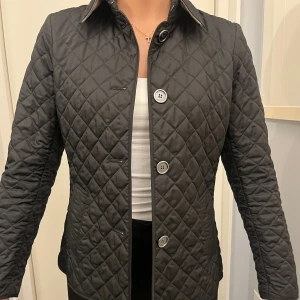 Svart quiltad jacka från Burberry - Snygg svart quiltad jacka från Burberry med klassiskt rutigt foder i beige, grått och rött. Jackan har knappar framtill, lång ärm och ett bälte bak med knappdetaljer. Perfekt för dig som gillar stilren design och ikoniska detaljer. Nypris 12000kr. Använd 2 gånger.