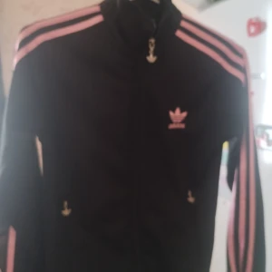 Svart Adidas track jacket med rosa ränder - Svart Adidas track jacket med rosa ränder längs ärmarna och rosa broderad logga både fram och bak. Jackan har dragkedja framtill och klassisk sportig look. Materialet är polyester och modellen är långärmad med normal passform.