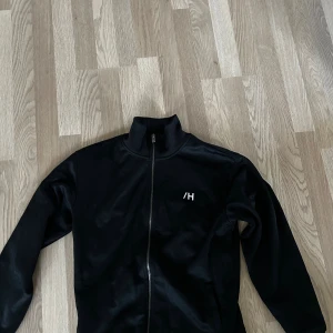 Svart zipjacka - Svart zipjacka med hög krage och vit /H-logga på bröstet. Jackan har hel dragkedja framtill och är tillverkad i mjukt syntetmaterial. Perfekt för en sportig och clean look.