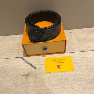 Svart Louis Vuitton rutigt skärp - Säljer ett stilrent Louis Vuitton bälte i svart skinn med klassiskt rutigt mönster och en stor LV-logga i svart metall som spänne. Bältet är brett och har en lyxig känsla, perfekt för dig som vill ha en statement-accessoar från ett ikoniskt märke.